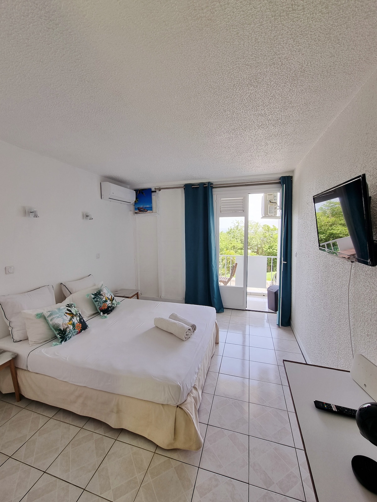 A bright sleeping area features a spacious bed dressed with light linens. Two decorative pillows add a touch of color. A flat-screen TV is mounted on the wall opposite a sliding door that opens to a balcony, enhancing natural light in the room.