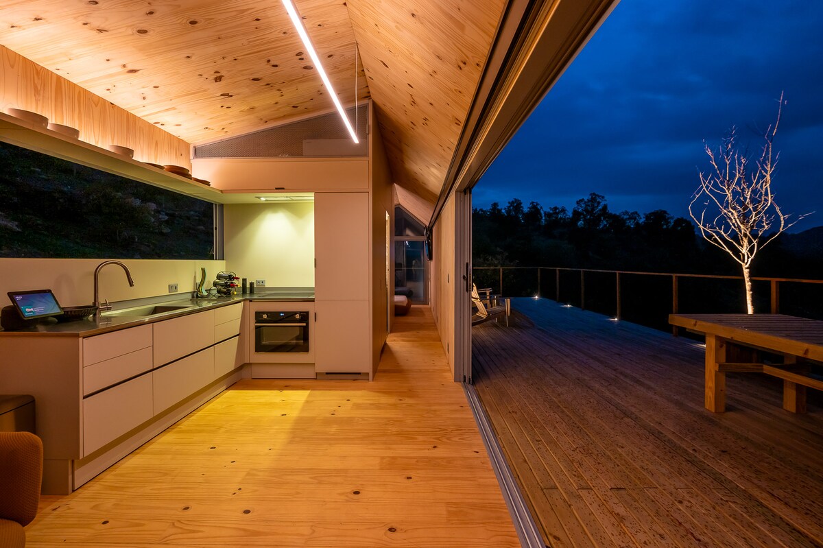 The interior space showcases an open kitchen area with modern appliances and light wood cabinetry. Large glass doors lead to an outdoor deck, enhancing the connection between indoor and outdoor living. Soft lighting creates a serene ambiance, while natural wood flooring adds warmth.