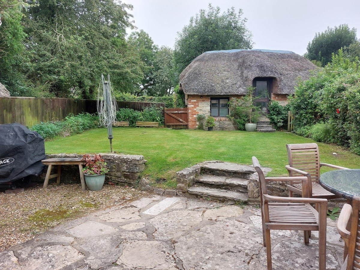 A well-maintained garden area is visible, featuring neatly trimmed grass and surrounded by lush greenery. Stone steps lead to a thatched-roof building, while patio furniture is arranged for outdoor relaxation. A barbecue unit is positioned nearby, adding to the outdoor amenities.