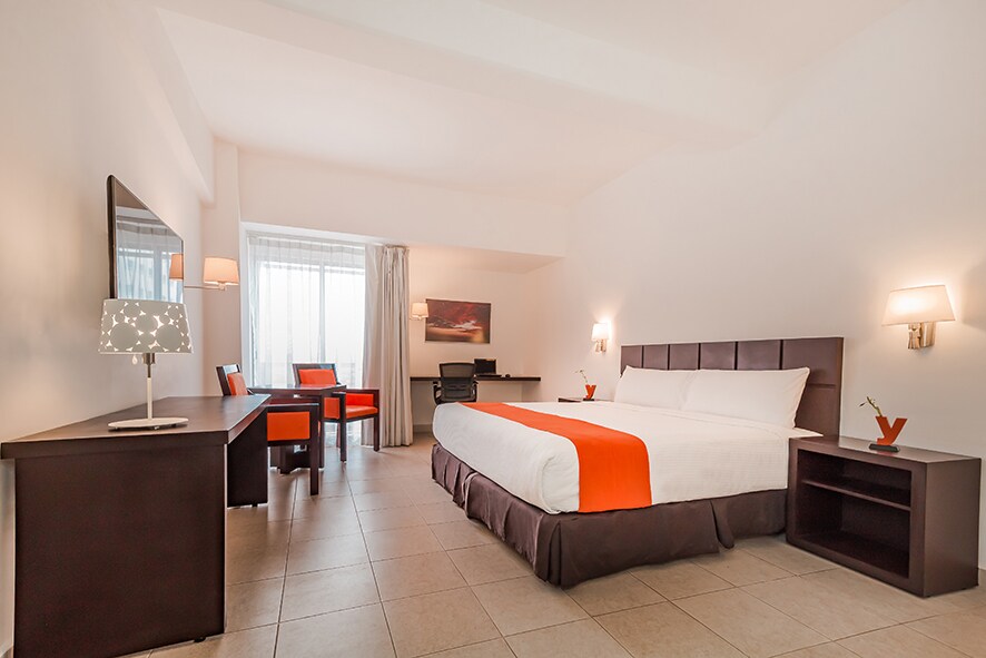 A light-filled guest room features a spacious bed with crisp white linens and a contrasting orange accent. A work desk and chair are positioned by the expansive window, alongside a modern lamp. Dark wood furniture adds a touch of sophistication to the neutral-toned space.