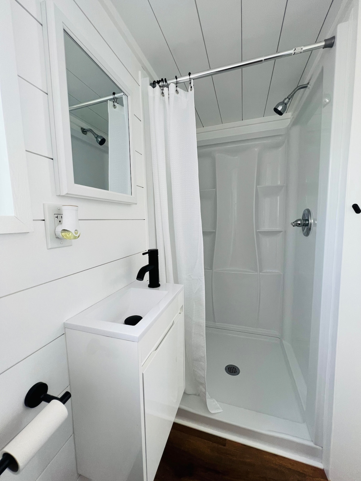 A modern bathroom features a walk-in shower with a transparent curtain and flush white walls. A compact sink is positioned beneath a mirrored cabinet, while a black faucet contrasts against the white fixtures. Natural wood flooring adds warmth to the space.