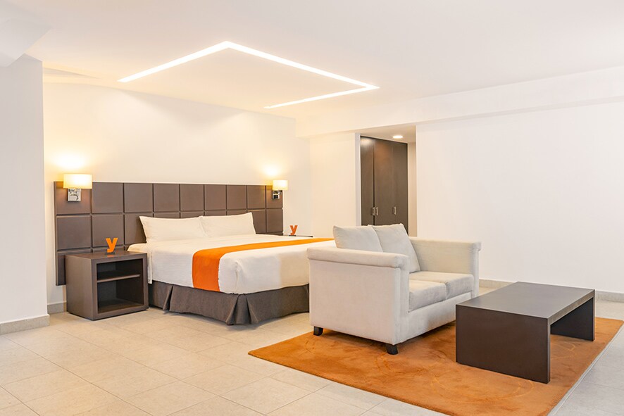 A modern guest room features a spacious bed with a warm orange accent, complemented by a comfortable seating area with a light-colored sofa and a dark coffee table. Soft lighting enhances the bright, neutral decor, creating an inviting space for relaxation.
