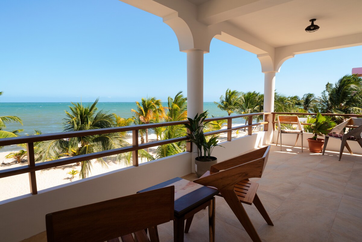 A spacious balcony provides panoramic views of the Caribbean Sea and sandy beach. Tropical palm trees sway in the gentle breeze, while wooden furniture invites relaxation. The area is bathed in natural light, highlighting the serene coastal atmosphere.