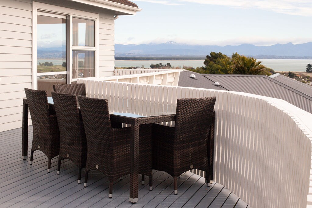 A dining set with a glass-top table and six dark wicker chairs is positioned on a spacious deck. A scenic view of Tasman Bay and distant mountains is visible in the background, framed by a white railing that offers a sense of openness.
