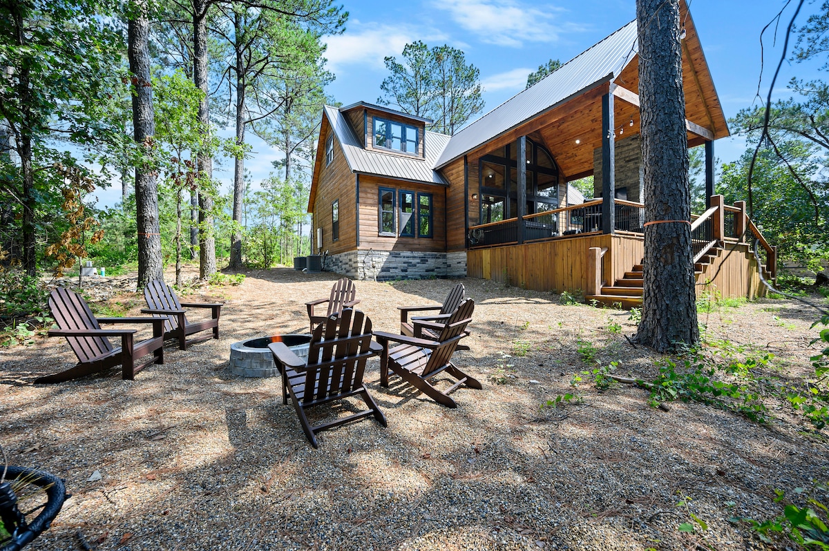 The exterior of the cabin is surrounded by a wooded area, featuring a spacious deck and seating arrangements. Several Adirondack chairs circle a fire pit, providing a cozy gathering space in a natural setting. The cabin's wooden facade contrasts with the lush greenery.