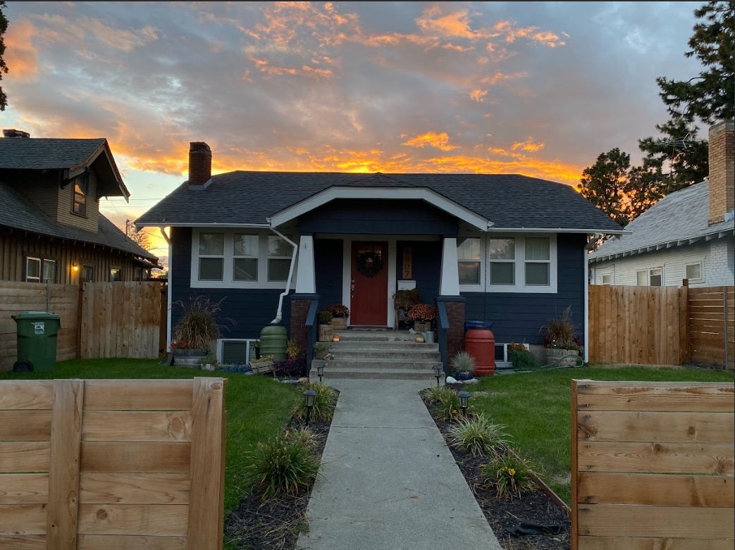 A blue Craftsman-style home with a welcoming entrance is highlighted against a vibrant sunset sky. The neatly paved pathway leads to the door, surrounded by landscaped greenery and flowerbeds. A fenced yard provides privacy, creating an inviting exterior atmosphere.