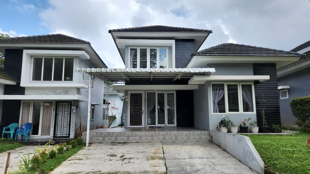 A modern two-story villa is presented with a sleek exterior design, featuring a combination of dark and light colors. A spacious porch is centered at the entrance, while large windows allow natural light to fill the interior. Neatly landscaped greenery flanks the sides of the villa.