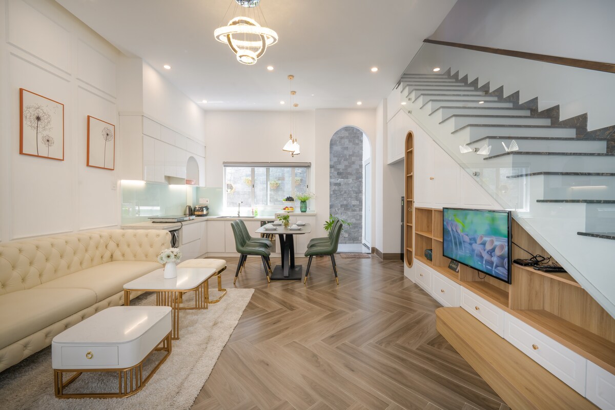A modern and spacious living area features a comfortable beige sofa, two white coffee tables, and a dining table set for four. Natural light fills the room through large windows. A staircase with glass railings leads to the upper floor, while a TV is mounted on a sleek wooden cabinet.