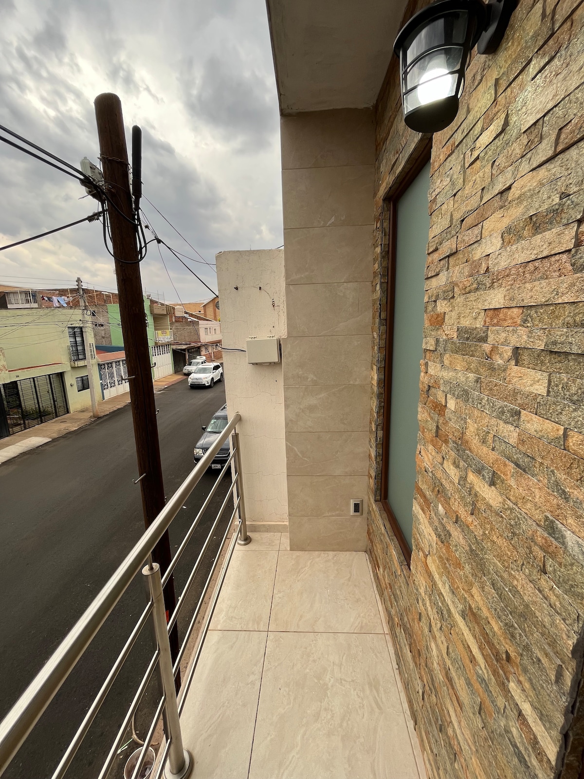 A balcony is presented with stone-clad walls and a metal railing. Neutral tile flooring extends outward, while a street view is visible beyond the railing. The setting features utility poles and parked cars, contributing to the urban landscape.