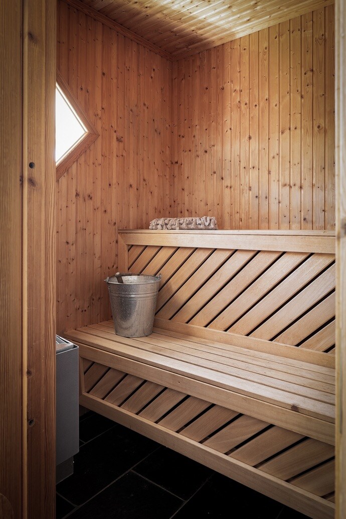 A wooden sauna is depicted with vertical paneling creating a warm ambiance. Two built-in benches provide seating along the walls. A small metal bucket is placed on one of the benches. Soft lighting enhances the natural wood tones throughout the space.