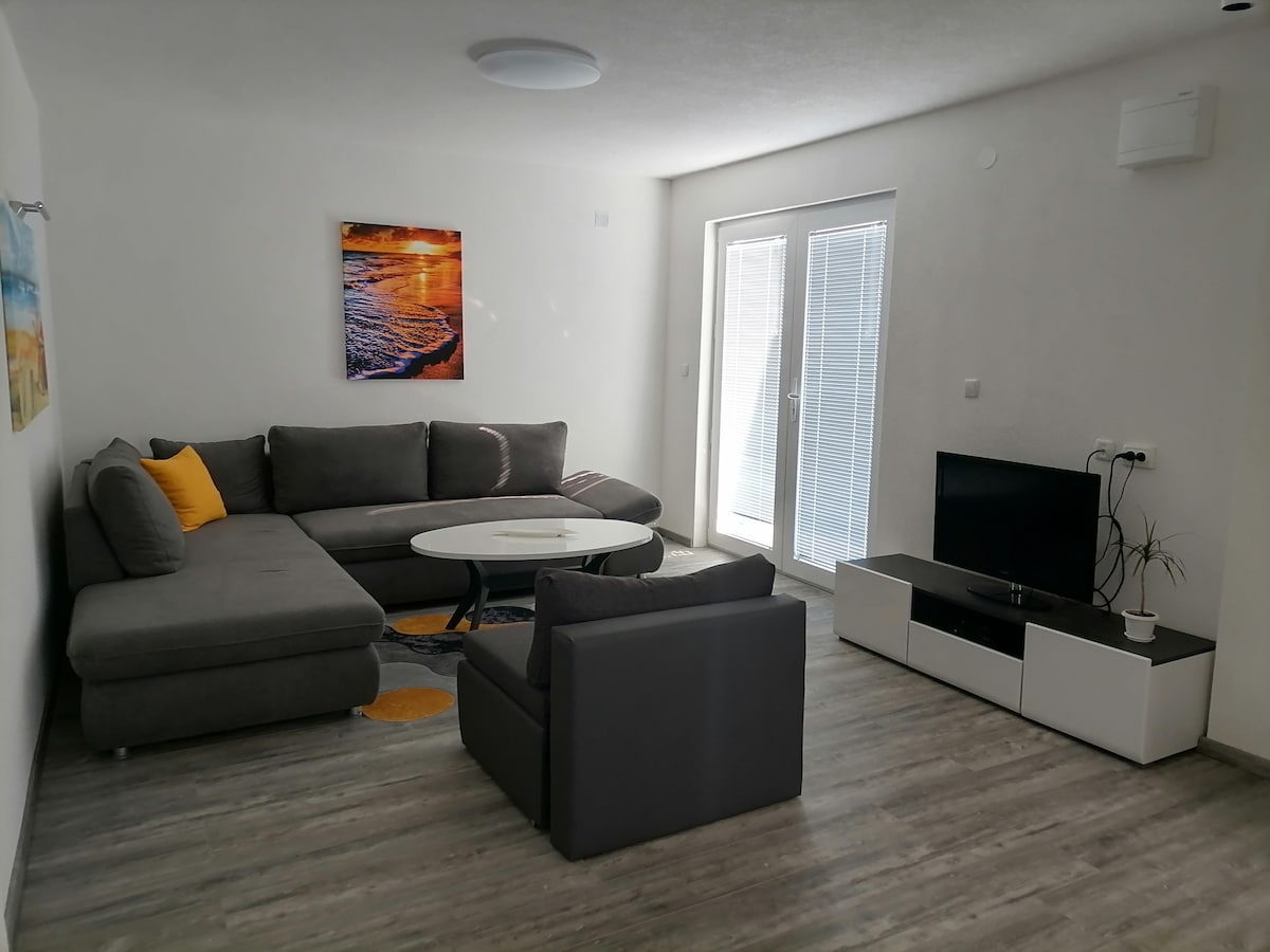 A spacious living area features a gray sectional sofa complemented by a round coffee table. Natural light enters through two glass doors. A simple television stand holds a television set, while a colorful wall art piece adds a touch of warmth to the neutral walls.