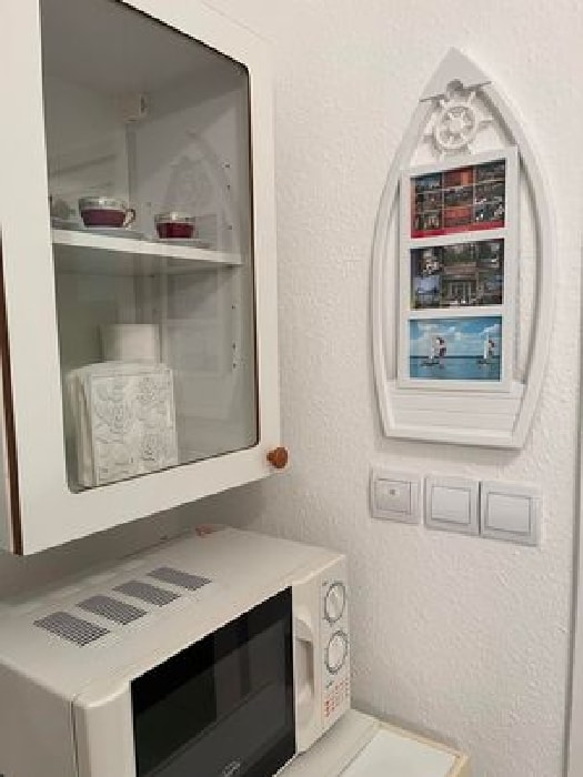 A compact kitchen area features a microwave below a display cabinet. The cabinet holds decorative items and glasses, while a framed collage of photos is mounted on the wall nearby. The overall space maintains a clean and organized appearance.