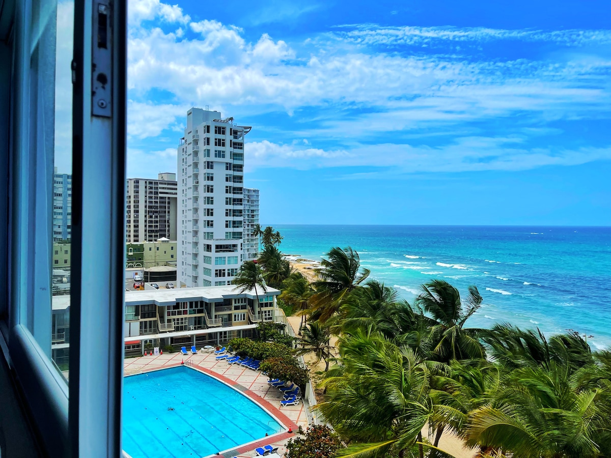 A panoramic view captures the ocean, showcasing gentle waves against sandy shores. In the foreground, a large swimming pool is framed by palm trees. The scene features tall buildings along the coastline, with vibrant blue skies enhancing the tranquil atmosphere.