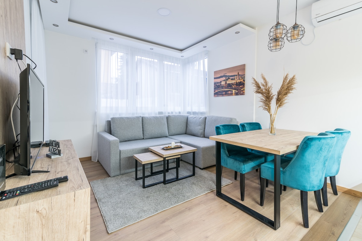 A modern living area features a spacious, light grey sectional sofa complemented by a small coffee table. A wooden dining table is surrounded by four teal chairs. Large windows allow natural light to fill the room, enhancing the neutral-toned decor.