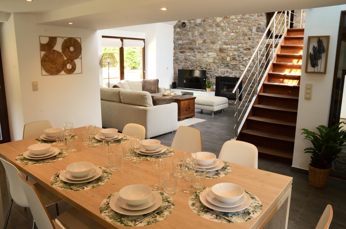 An open-plan living and dining area features a wooden dining table set for a meal, with patterned placemats and glassware. A stone wall adds texture, while a staircase leads to the upper level. A cozy seating arrangement includes a large couch facing a television.