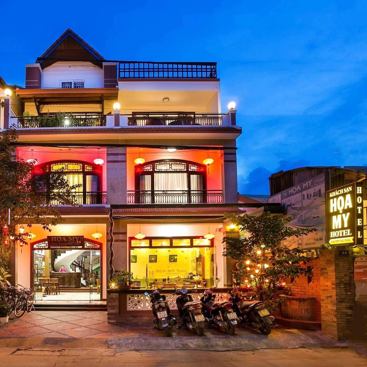 The exterior of the building is highlighted by warm lights, creating a welcoming ambiance. A spacious entrance leads into the hotel, flanked by parked motorcycles and bicycles. The three-story structure features balcony spaces and large windows, enhancing the inviting atmosphere.