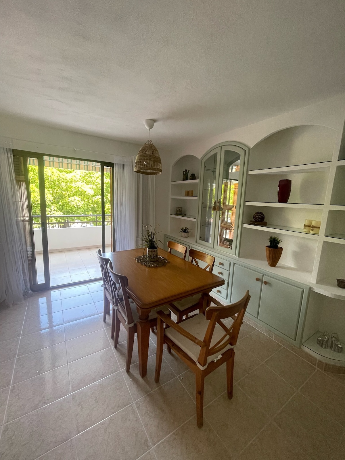 A dining area features a wooden table surrounded by six chairs. Natural light streams through sliding glass doors that lead to a balcony, enhancing the space’s brightness. A built-in cabinet and shelves display decorative items, contributing to the room's welcoming ambiance.
