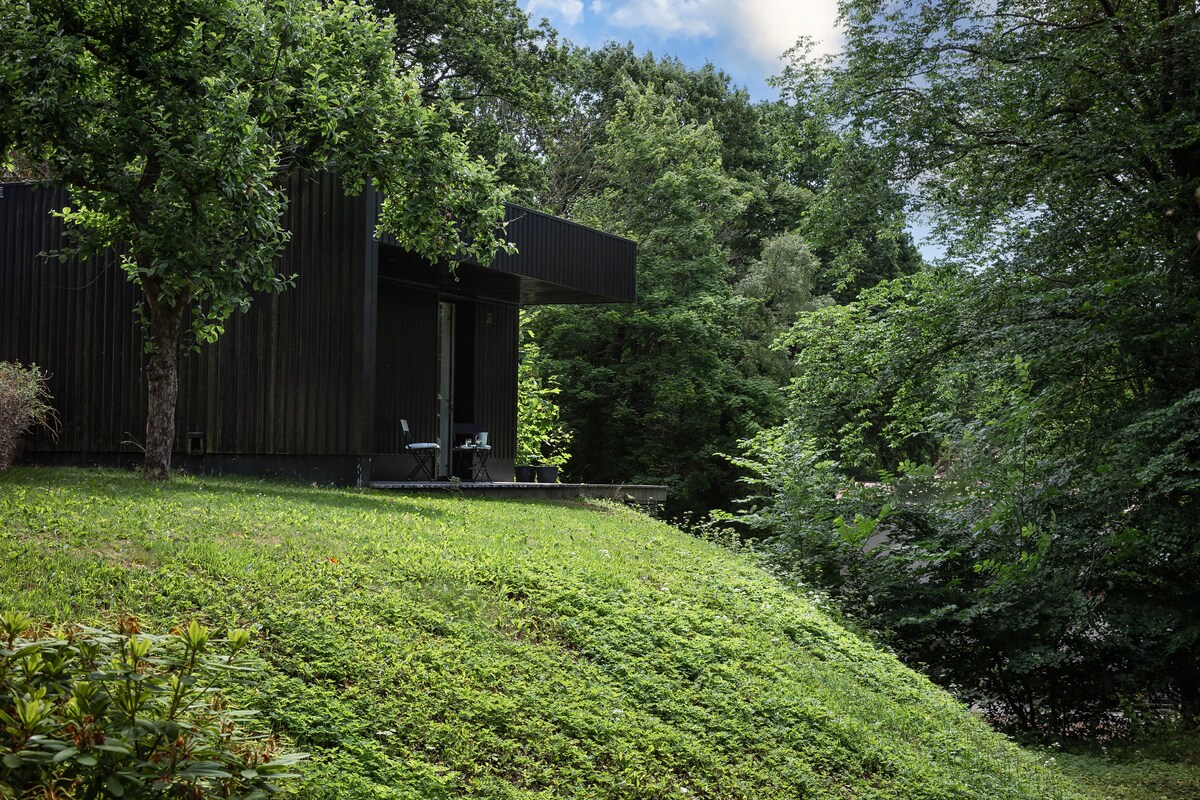 A modern black structure is nestled among lush greenery, perched on a grassy slope. Two chairs are positioned on the patio, offering a peaceful view of the surrounding trees and shrubs. The natural landscape creates a serene setting, ideal for relaxation.