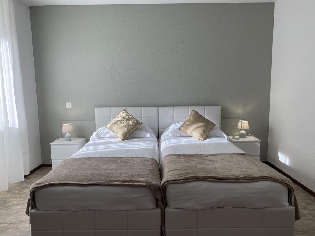 A well-lit sleeping area features two spacious beds with neutral bedding, complemented by decorative pillows. Soft white bedside tables with lamps are positioned on either side, enhancing the serene atmosphere. Natural light enters through a large window, with sheer curtains allowing for privacy.