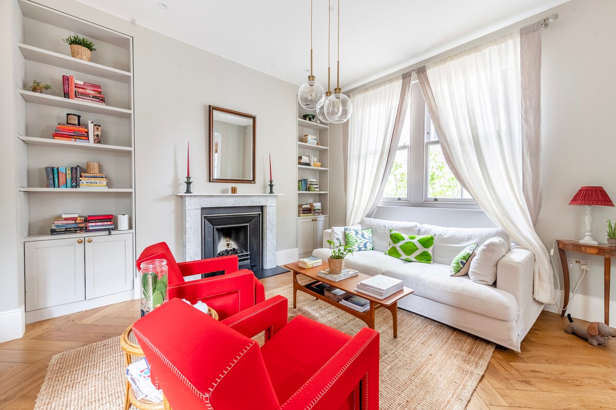 A bright living area features a white sofa accompanied by two red chairs, positioned on a woven rug. A sleek coffee table is centered in front of the seating. The walls are lined with shelf units filled with books and decorative items, while natural light filters through sheer curtains.