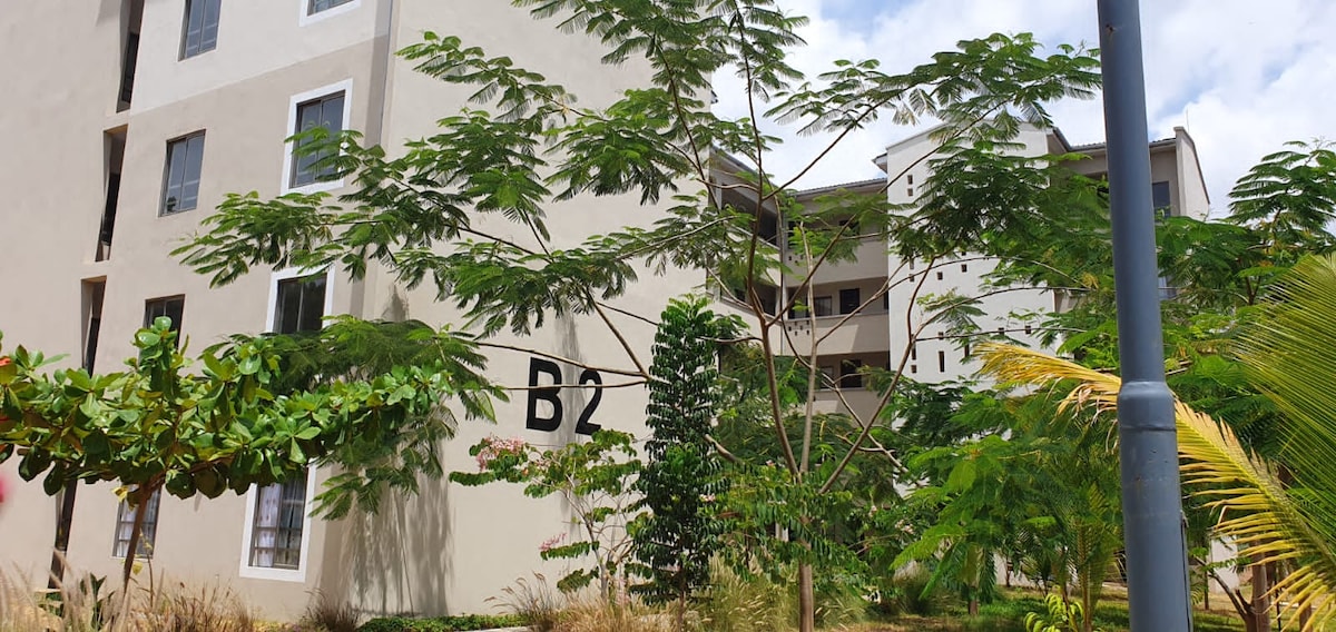 A multi-story building is visible, marked with the number 'B2.' Lush greenery, including palm trees and other foliage, surrounds the structure, complementing its modern façade. Soft light filters through the leaves, and a clear sky is visible above.