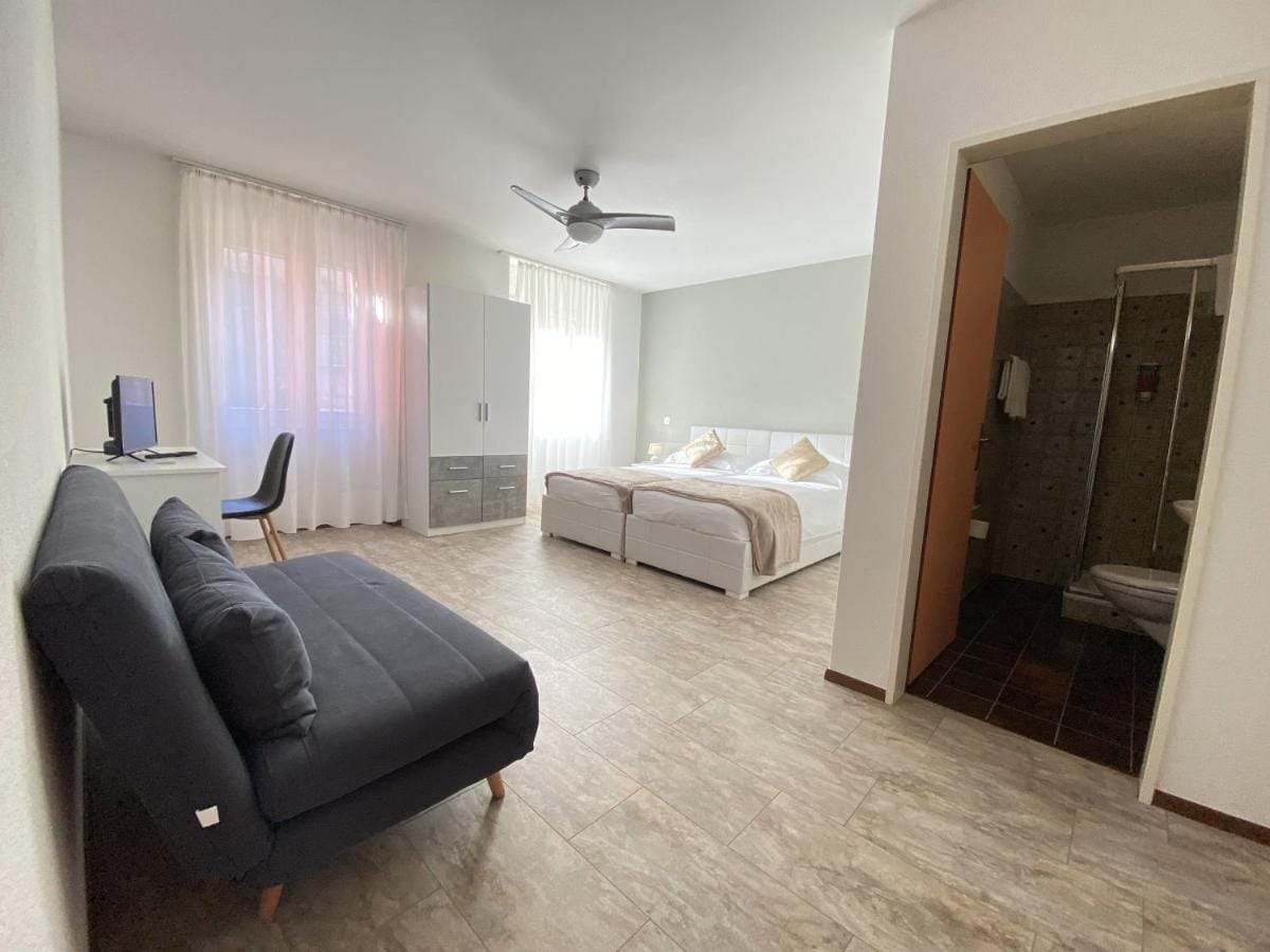 A bright and spacious guest room features two beds arranged together. Natural light enters through sheer curtains, illuminating the room. A modern desk and chair are positioned near a wall-mounted television, while a comfortable sofa enhances the seating area. A bathroom is accessible through a door.