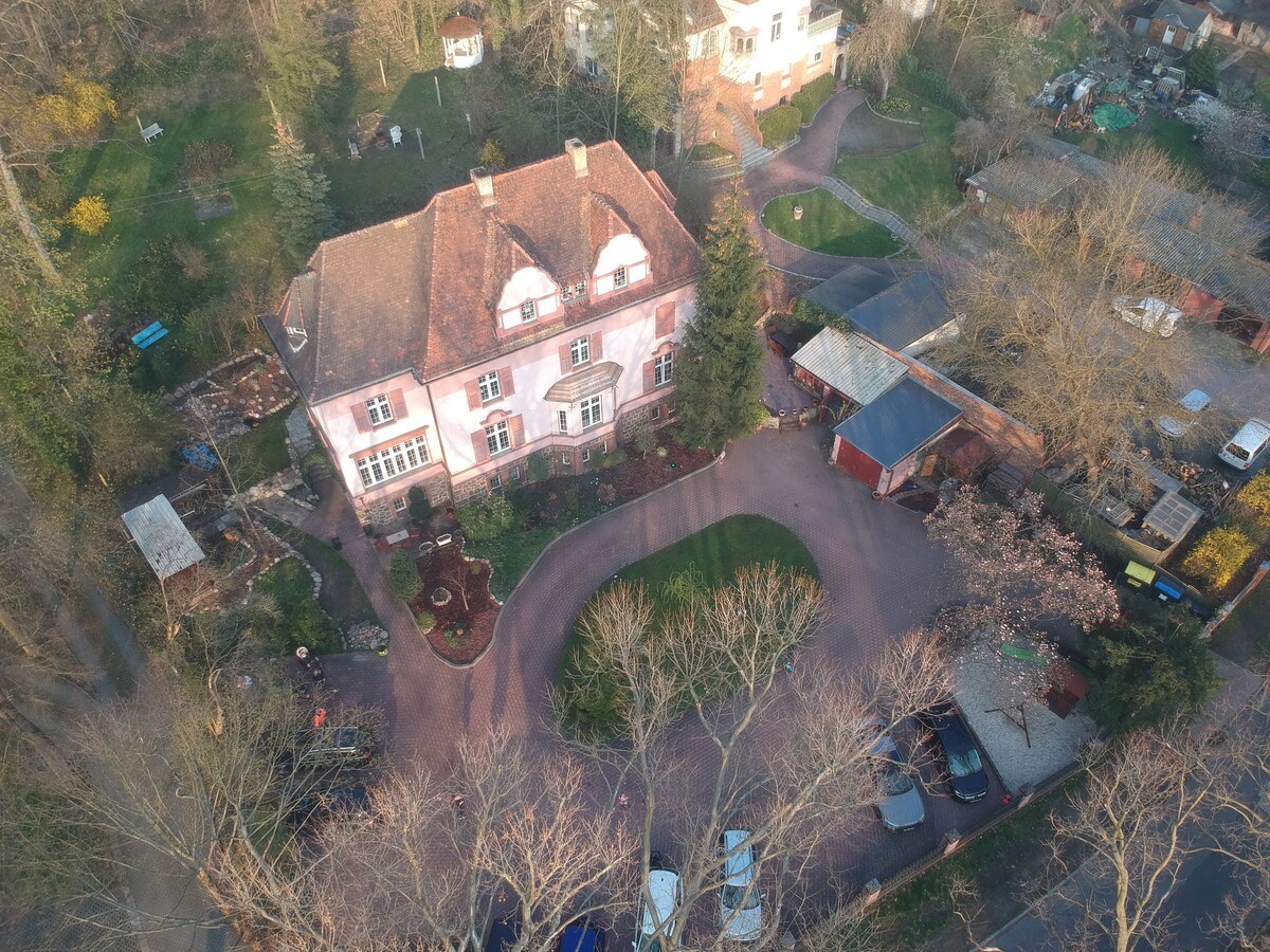 An aerial view captures a charming pink villa set amidst lush greenery. The property features a circular driveway, landscaped gardens, and various trees. A mix of parked cars and nearby buildings is visible, showcasing a serene and spacious outdoor setting.