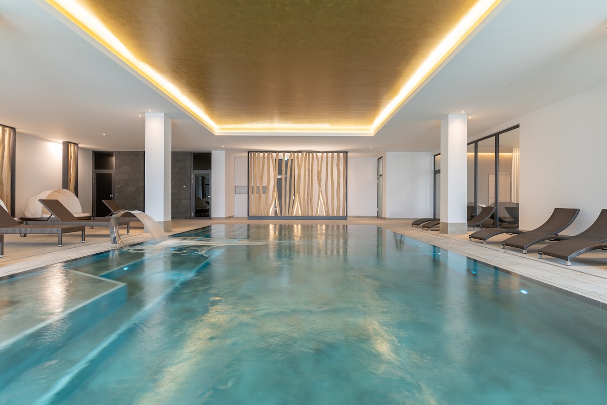 An indoor wellness area features a serene swimming pool surrounded by sleek lounge chairs. Soft lighting reflects off the water's surface, and large glass panels offer a glimpse of the room's modern design and tranquil atmosphere.