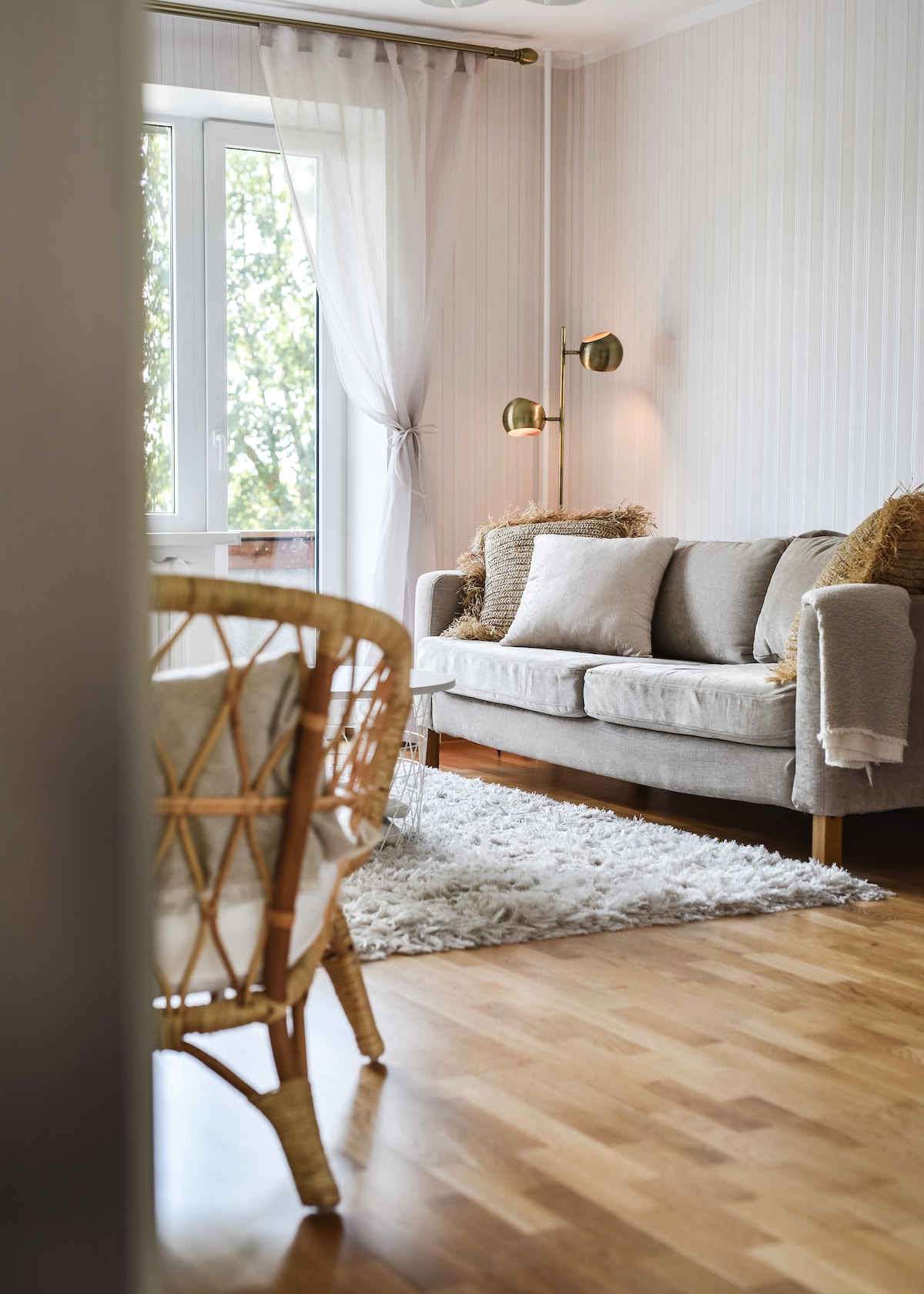 A living area is presented with a light grey sofa adorned with soft pillows. A cozy rug is placed on polished wooden flooring. Natural light filters through a large window with sheer curtains, creating a relaxing atmosphere. A comfortable armchair complements the seating arrangement.