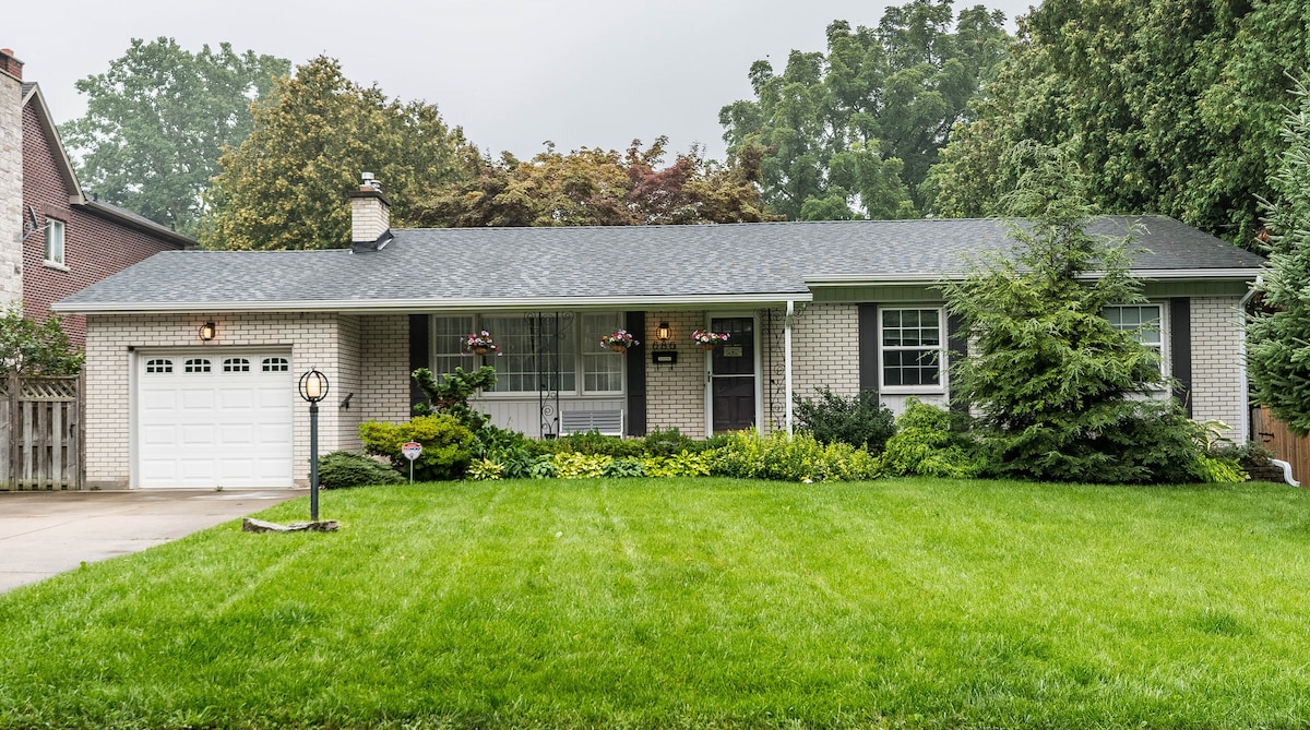 The front exterior of the home is showcased, featuring a well-maintained lawn and lush greenery. Flower baskets hang from the windows, and a light fixture stands near the entrance. A spacious driveway is visible, accommodating multiple vehicles, under a canopy of trees.