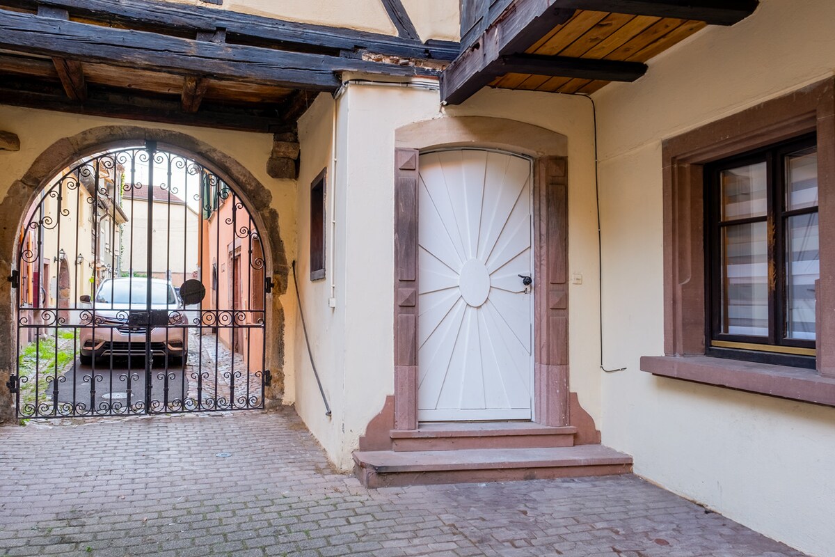 An arched stone gate opens to a charming courtyard, featuring a sleek car parked on a cobblestone pathway. The white door, decorated with a circular design, leads indoors. Nearby, a window offers a glimpse of the interior, framed by rustic building details.