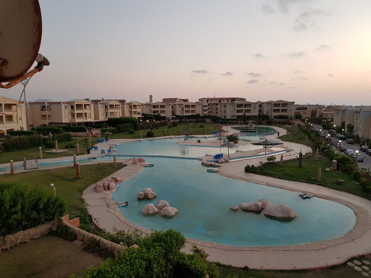 An expansive swimming area features clear water surrounded by landscaped greenery and rocky formations. Several lounge chairs are positioned by the water's edge. Residential buildings are visible in the background, with a peaceful sunset enhancing the serene atmosphere.