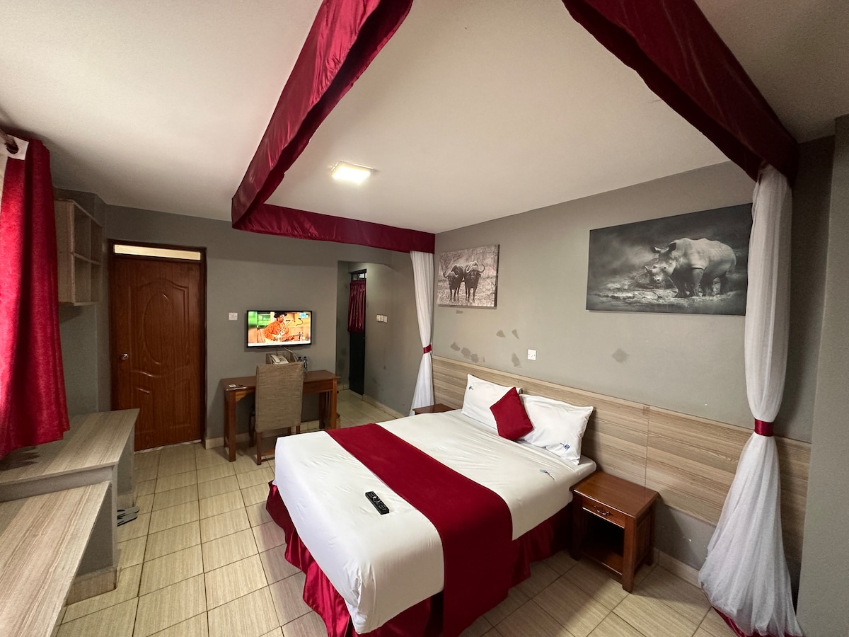 A well-appointed room is showcased with a spacious bed dressed in crisp white linens and a red decorative runner. A small desk is positioned near the window, and a 43'' TV is mounted on the wall. Soft red curtains frame the windows, adding warmth to the setting.