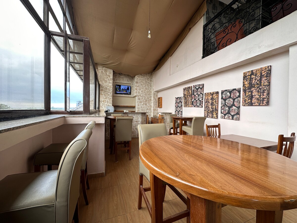A modern dining area is emphasized by a spacious wooden table surrounded by upholstered chairs. Large windows provide natural light and views, while decorative wall art adds visual interest. A flat-screen TV is mounted near the bar area, creating a relaxed social atmosphere.
