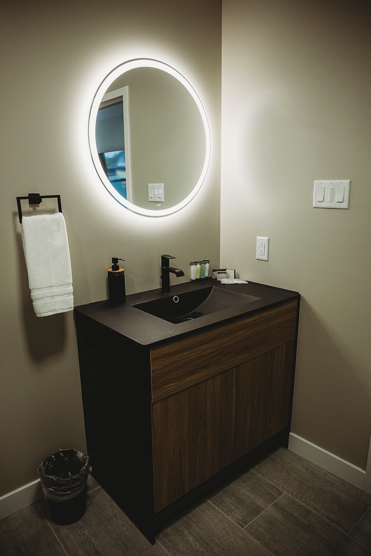 A modern bathroom vanity is equipped with a circular mirror lit by an integrated light. The dark cabinet features a sleek sink and is complemented by personal care items nearby. A neatly hung towel and wastebasket complete the functional design.