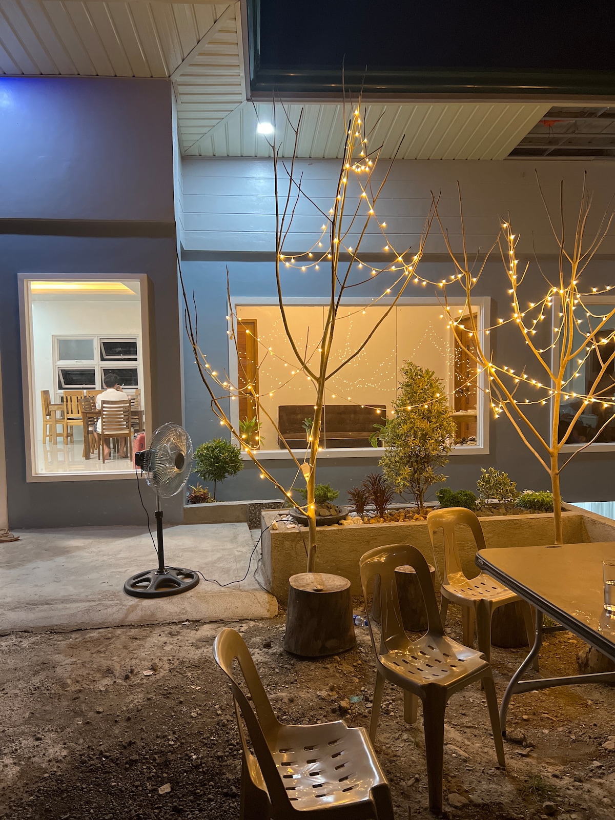 An outdoor space features a simple table and several chairs arranged around it. Two trees are adorned with twinkling fairy lights, adding a soft glow. A fan stands nearby, while a window offers a glimpse into the well-lit interior of the house.