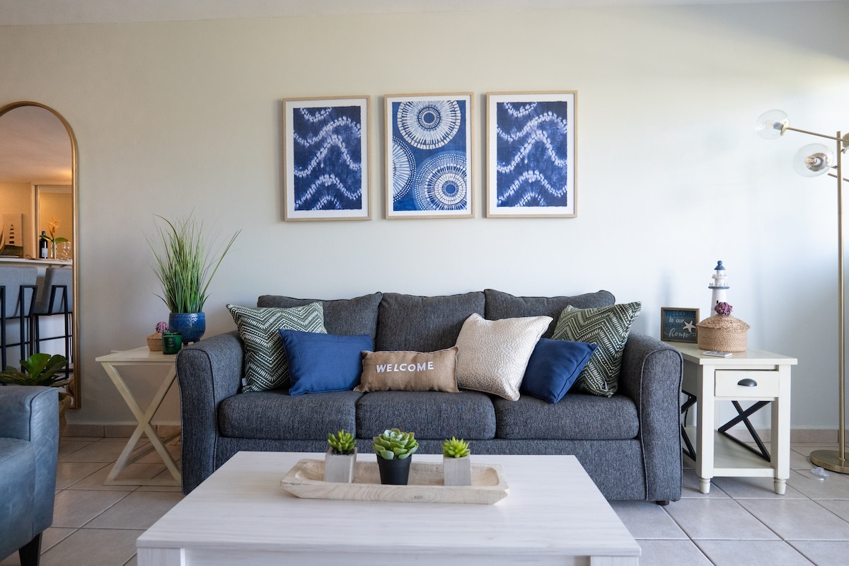 A comfortable living area is presented with a gray sofa adorned with decorative pillows in shades of blue and green. A welcoming pillow is centered on the sofa. Above, three framed artworks with blue patterns are displayed. A light wooden coffee table holds small potted plants.