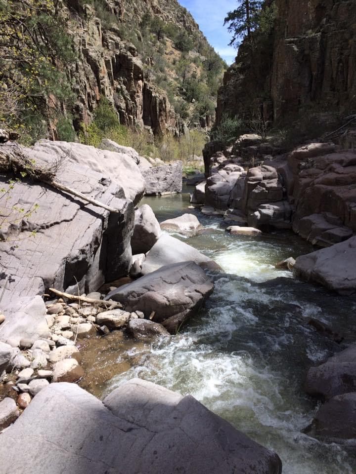 A narrow river flows gently through rocky terrain, flanked by steep canyon walls adorned with greenery. Smooth stones line the riverbed, while sunlight reflects off the water, creating a serene natural setting perfect for exploration.