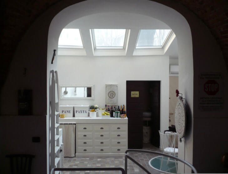 An airy kitchen area is visible, featuring a white cabinetry and a center island. Skylights allow natural light to fill the space, enhancing the warm tones of the wooden accents. Bottled beverages and fresh produce are arranged on the counters, indicating a well-equipped area for meal preparation.