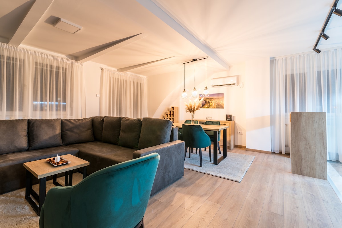 A well-lit living space is highlighted by large windows covered with sheer curtains. A sectional sofa in a comfortable gray tone complements a wooden coffee table. A dining area with a table and pendant lights completes the space, enhancing its modern design.