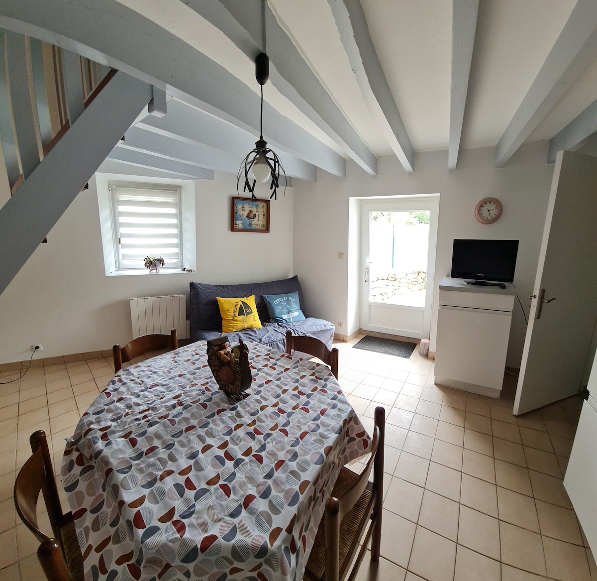 A bright living area features a dining table with a patterned tablecloth and four wooden chairs. A cozy sofa is positioned against the wall, with a television nearby. The space is complemented by white walls and wooden beams overhead, offering an inviting ambiance.