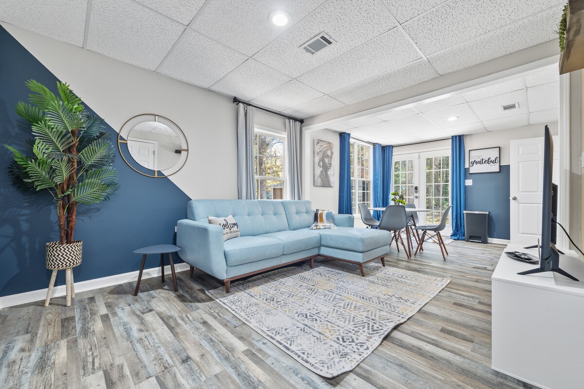 A living area features a light blue couch positioned against a two-tone wall, complemented by a round mirror. Natural light fills the space from large windows with blue curtains, while a patterned rug and a small table add to the welcoming atmosphere.