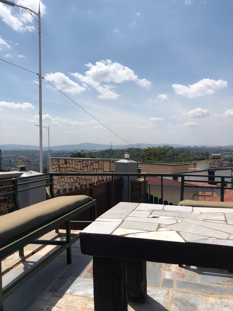 A rooftop space is featured with a comfortable seating area. The surface is made of stone, and the view includes distant hills under a clear sky with scattered clouds. The railing surrounds the area, providing safety while enjoying the landscape.