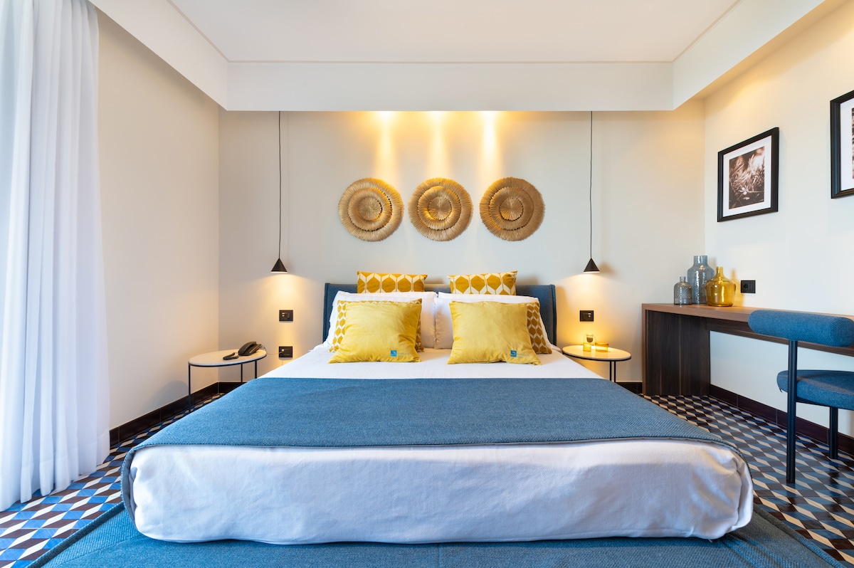 A modern bedroom features a spacious bed adorned with blue and yellow pillows. Three decorative woven wall hangings add texture above the bed. A small desk and chair are positioned against the wall, complemented by a geometric patterned floor.