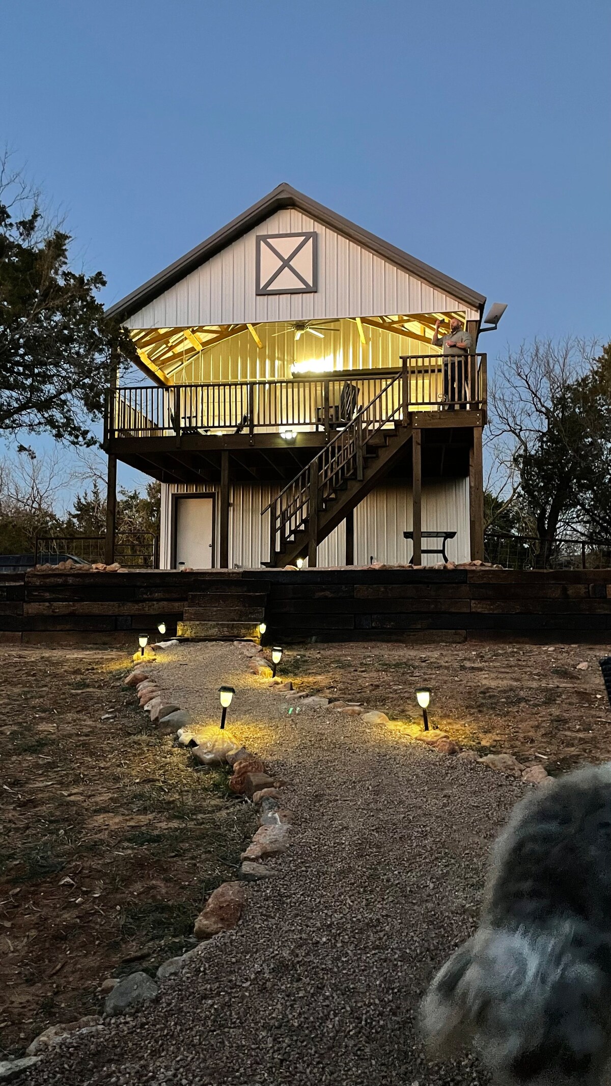 The exterior of the two-story house is illuminated by warm lights, enhancing its inviting presence. A well-defined path, lined with soft lighting, leads to the entrance. Surrounding greenery adds to the home's natural setting, creating a serene ambiance at dusk.