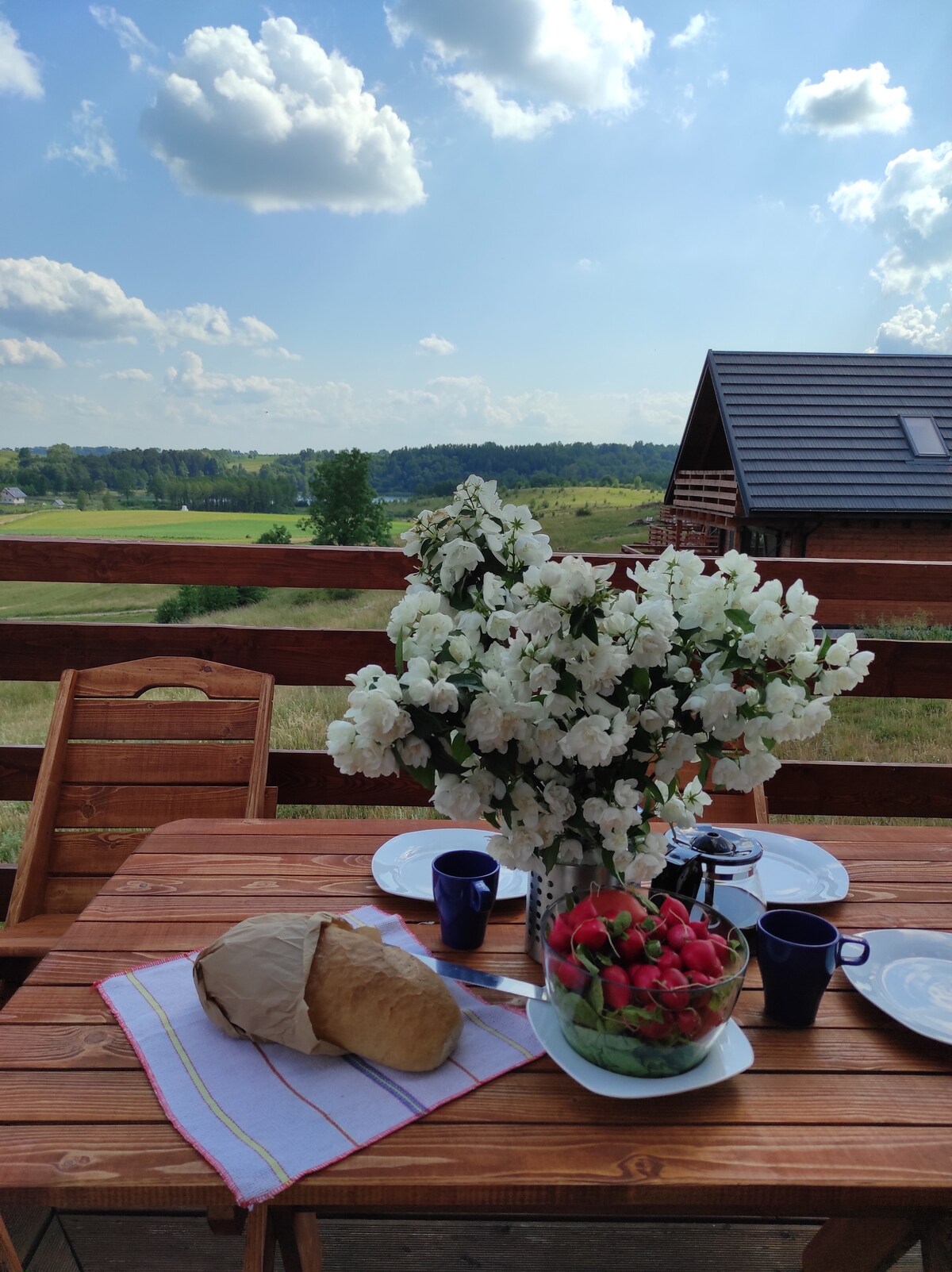 A wooden outdoor table is set for a meal, featuring dishes of strawberries and bread. A bouquet of white flowers adds a fresh touch, while a backdrop of rolling green hills and a clear blue sky enhances the peaceful setting.
