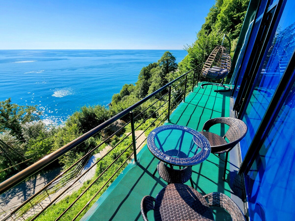 A spacious balcony offers stunning views of the ocean, with sunlight reflecting on the water's surface. Two woven chairs and a small round table are positioned on a green mat, surrounded by lush greenery. The large glass windows of the building enhance the connection to the outdoors.