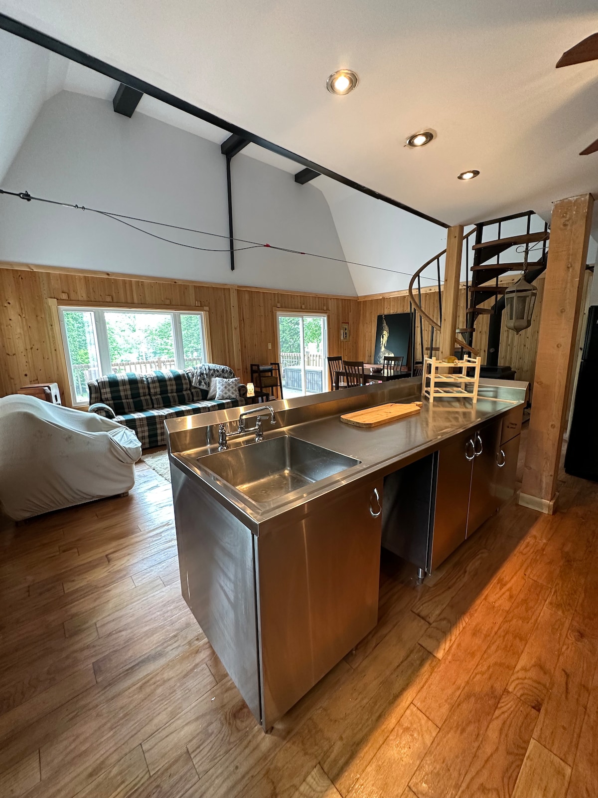 A modern kitchen features a stainless steel island with a wooden cutting board on the counter. Overhead lighting highlights the open space, which connects to a cozy seating area with a plaid couch and large windows allowing natural light to fill the room.