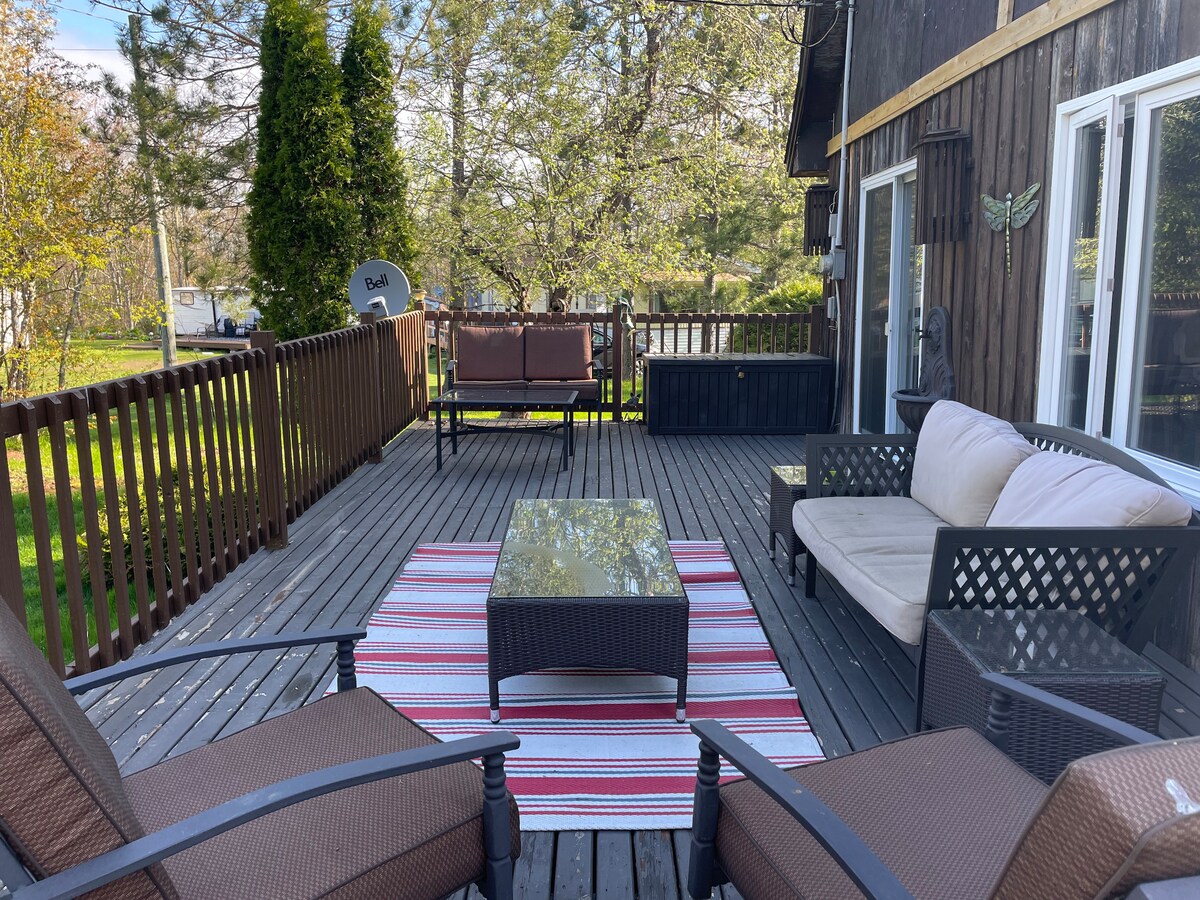 An outdoor deck area features a seating arrangement with a coffee table and multiple comfortable chairs. A striped outdoor rug adds a pop of color. Lush greenery surrounds the space, creating a relaxing atmosphere for enjoying the fresh air.