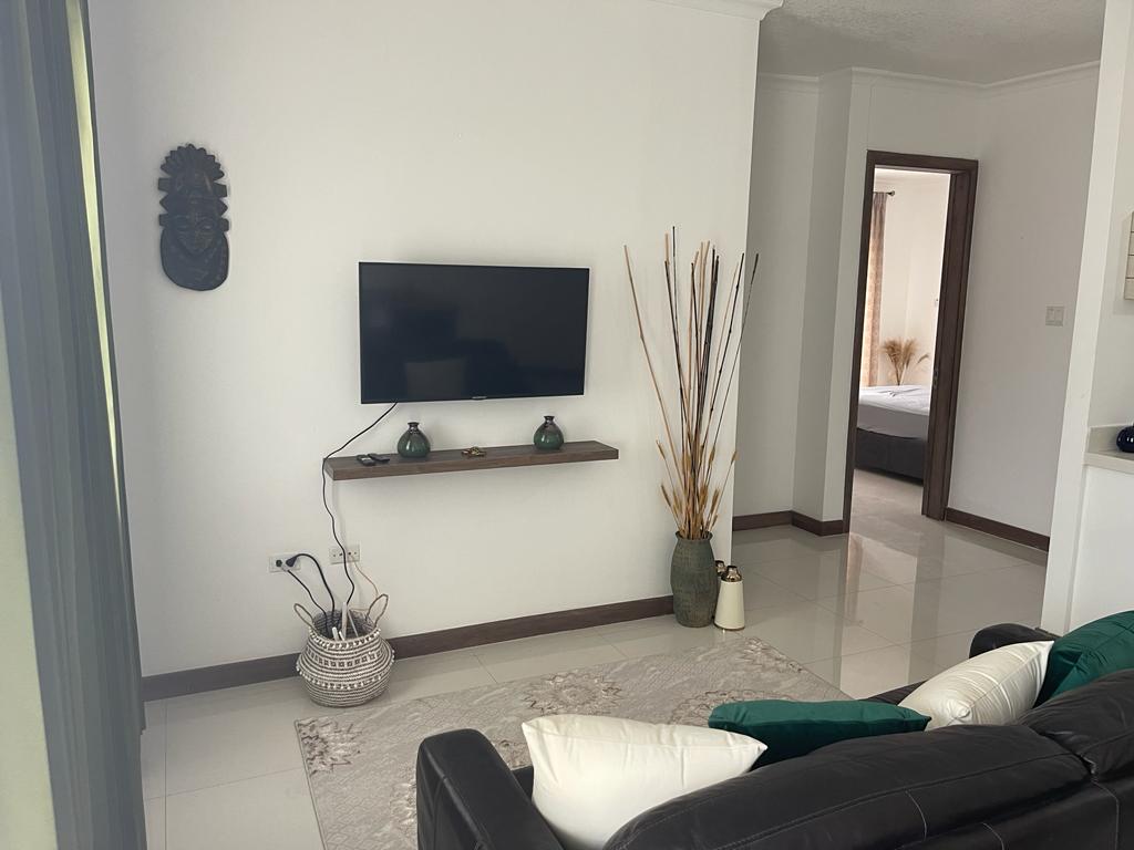 A contemporary living area features a mounted flat-screen television on a minimalist shelf. Decorative vases and plants add a touch of style beside a comfortable black sofa adorned with textured throw pillows. The space is illuminated by natural light, inviting a sense of relaxation.